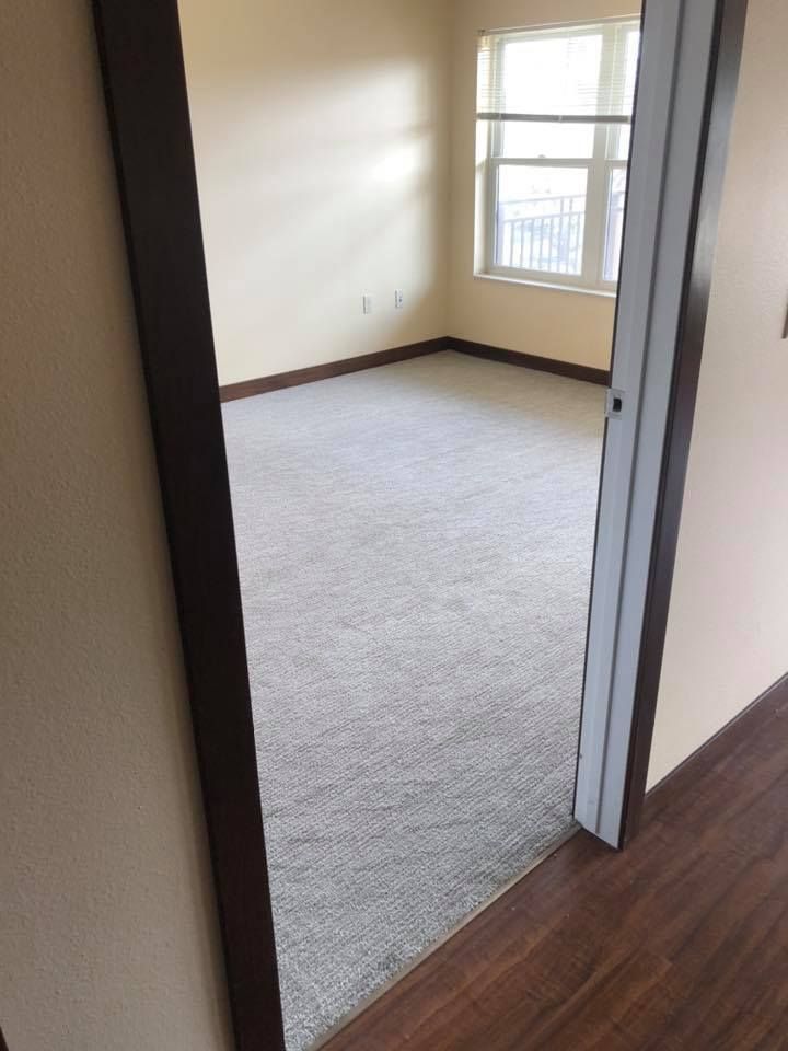 Empty carpeted bedroom with beige walls and a window, viewed from the doorway.