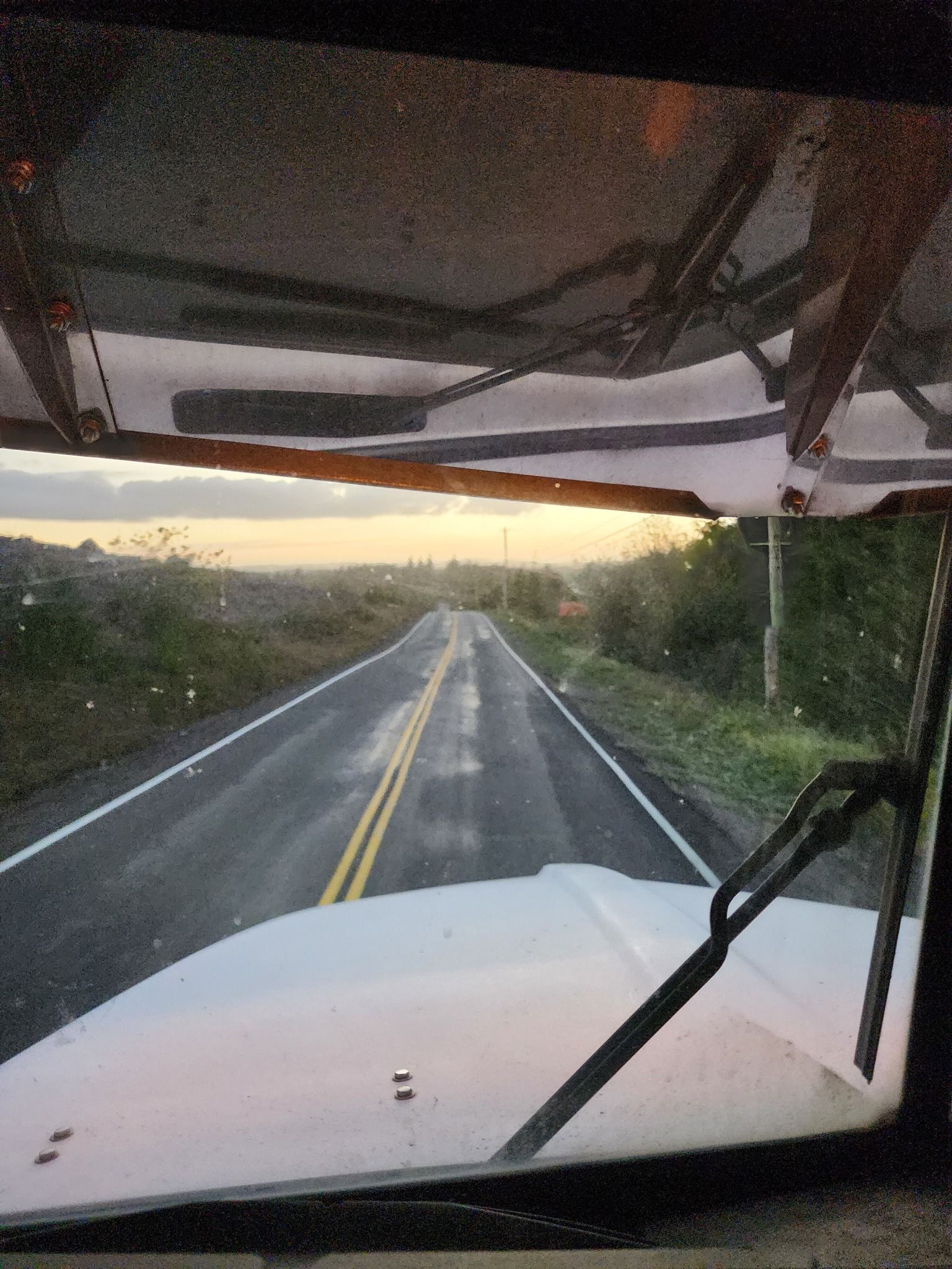 View from a truck cab driving on a wet two-lane road at sunrise, with windshield wipers visible.