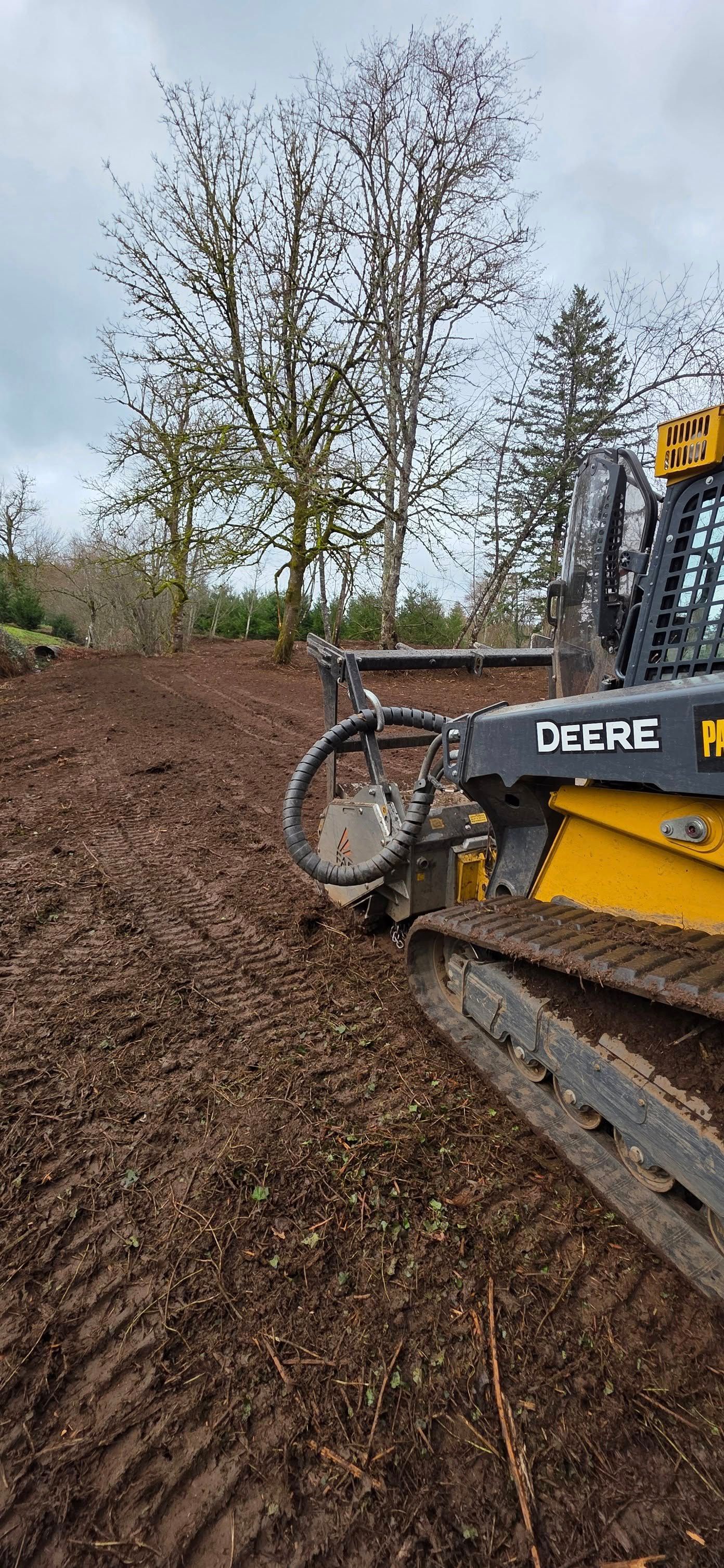 Yellow Deere excavator arm clearing muddy ground near leafless trees in a field