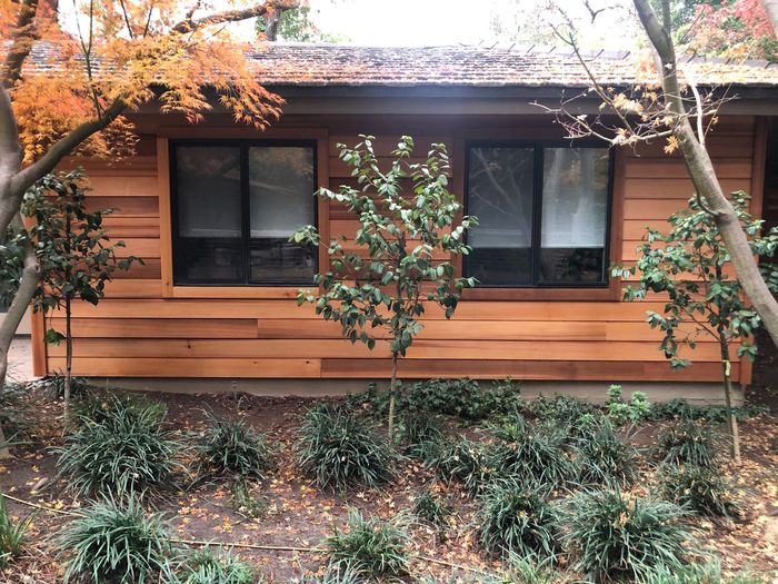 a house with wooden siding and a roof is surrounded by trees and bushes .