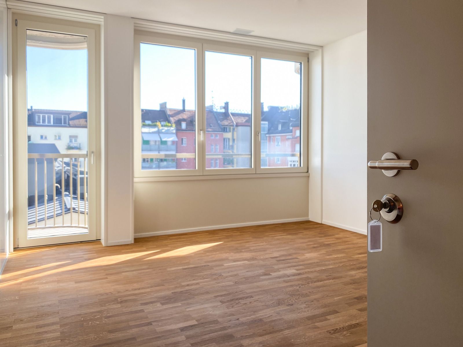 Empty sunlit room with a balcony, large windows overlooking colorful buildings, and a door
partially