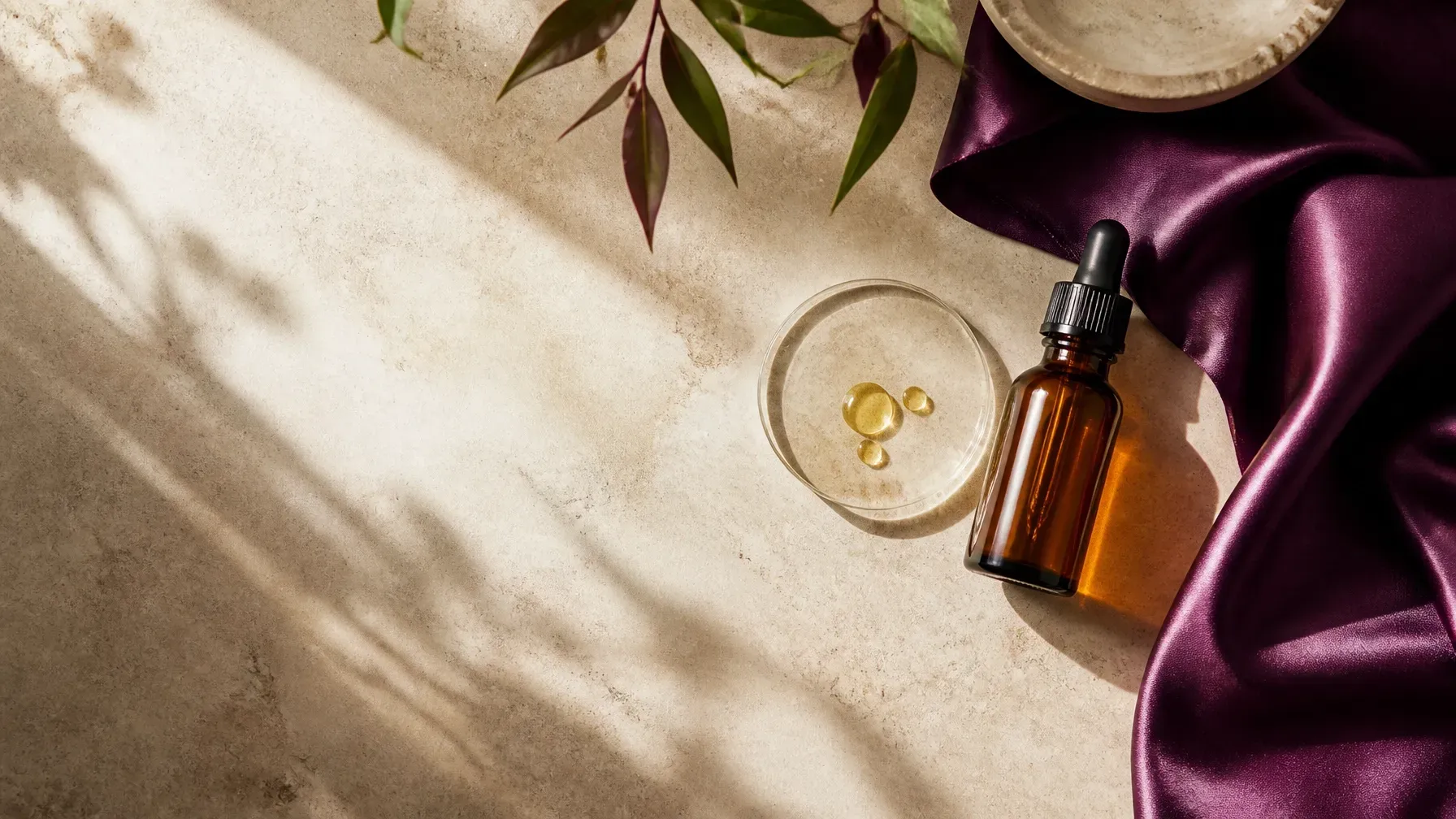 Amber bottle and glass dish on beige fabric with purple satin and dried leaves, lit by soft sunlight