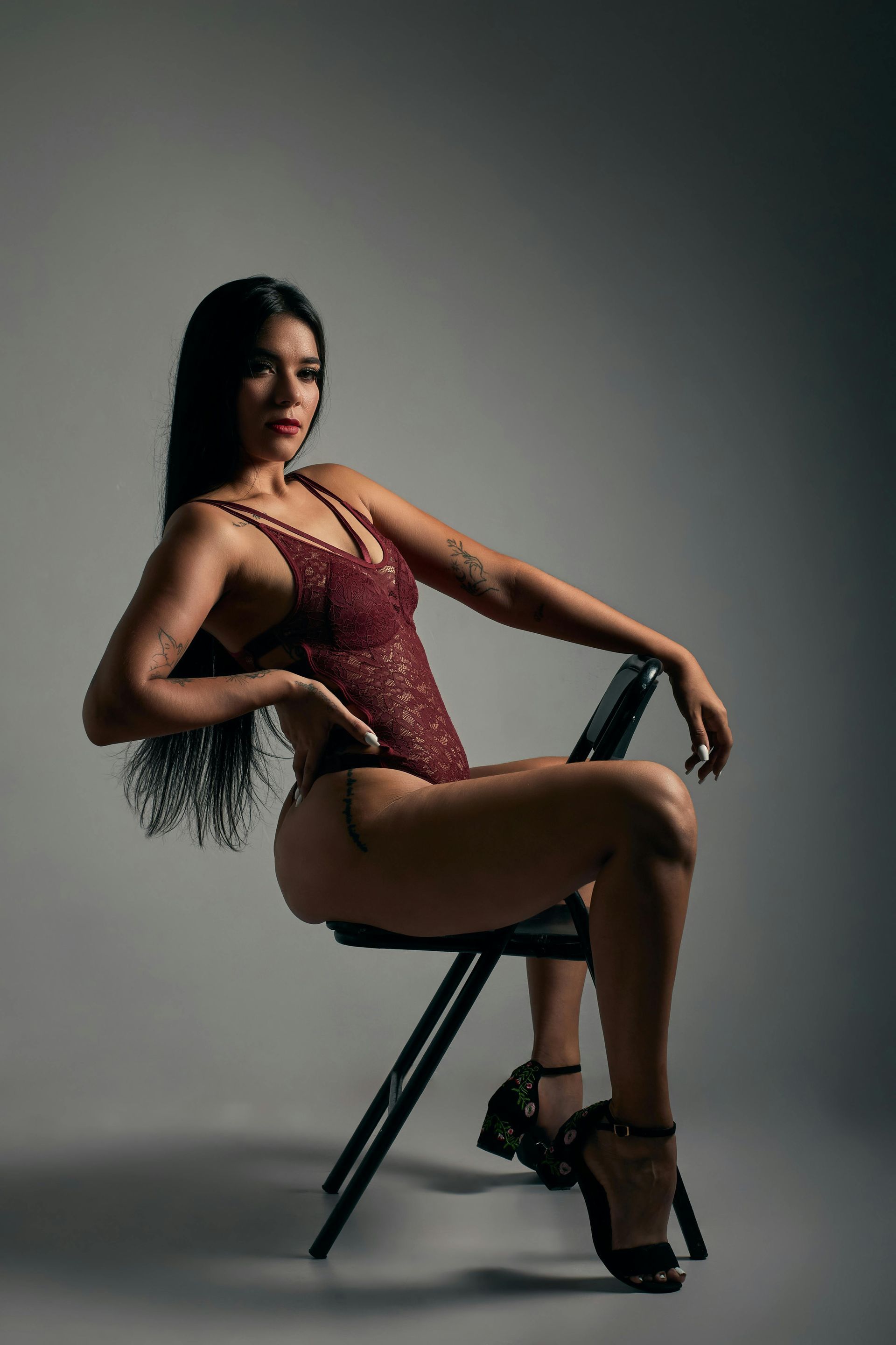 Woman posing on a chair in a studio, wearing a burgundy bodysuit and black heels.