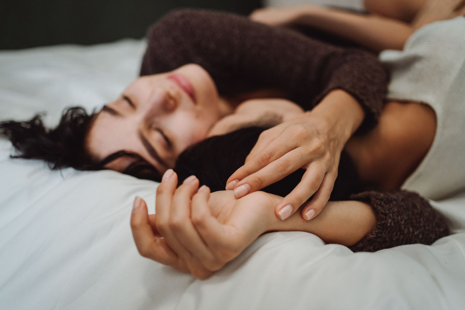 Person lying on a bed, eyes closed, gently holding another person’s hand in a close embrace