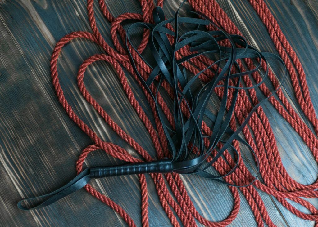 A bunch of red ropes and a black whip are laying on a wooden table.