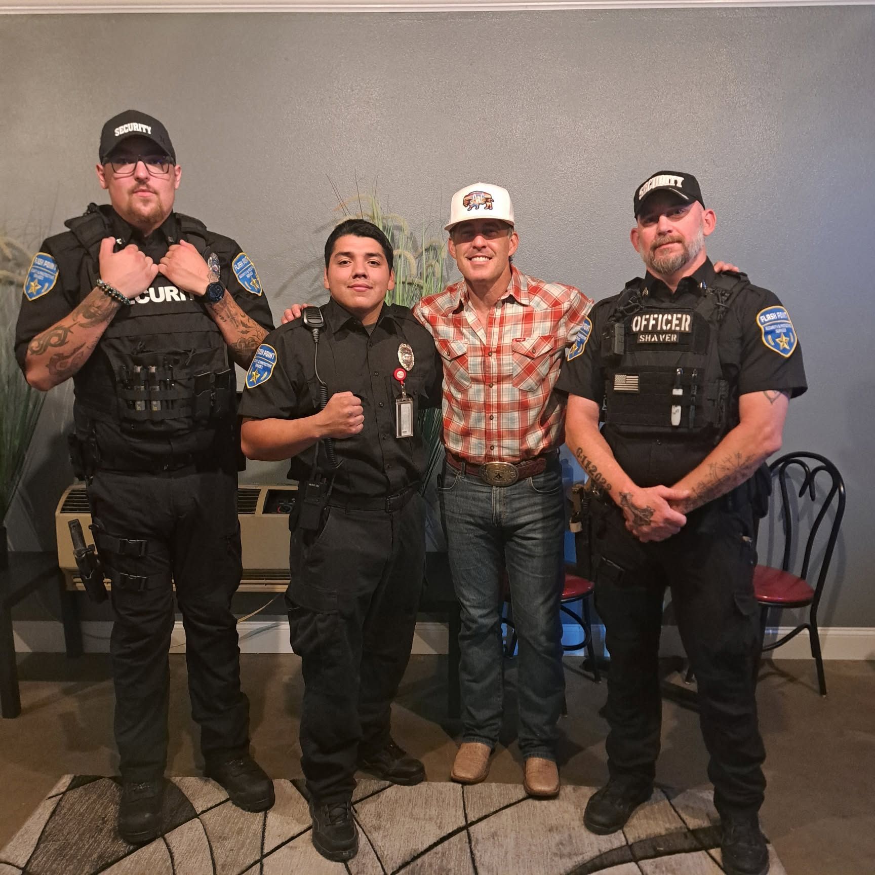 Four people posing indoors, including two police officers, with one man in a plaid shirt and cowboy hat.