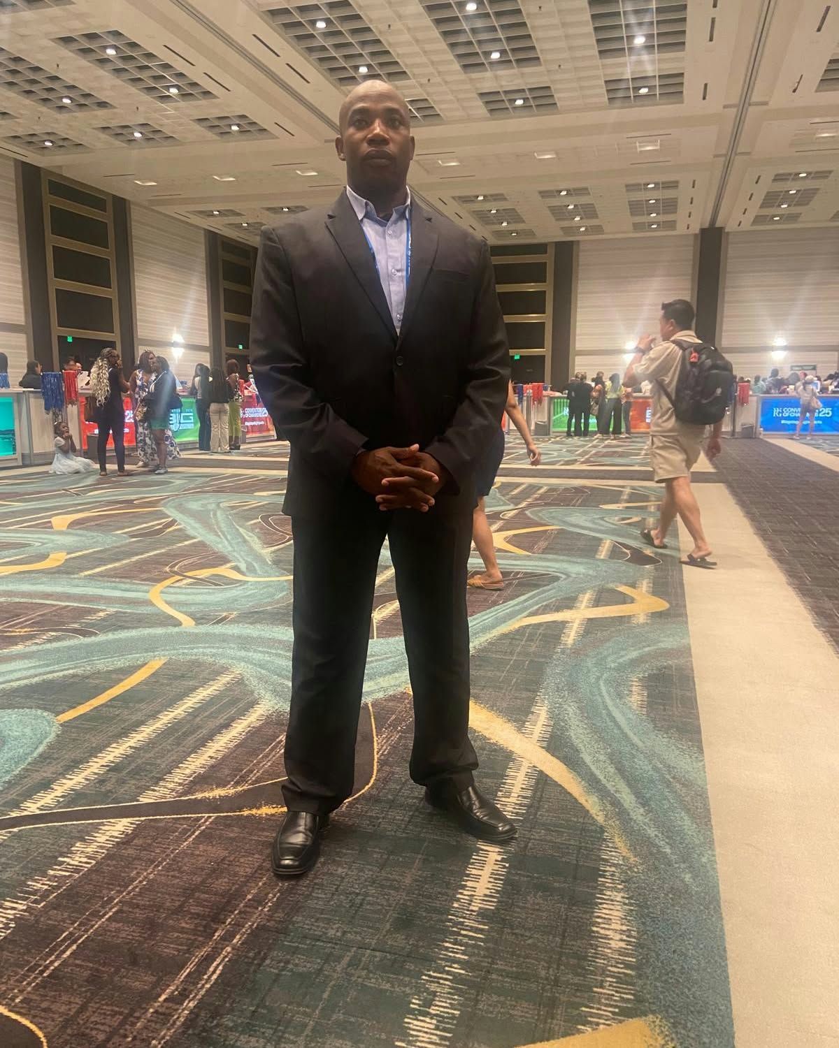 Man in a dark suit standing in a hotel ballroom with patterned carpet and people in the background
