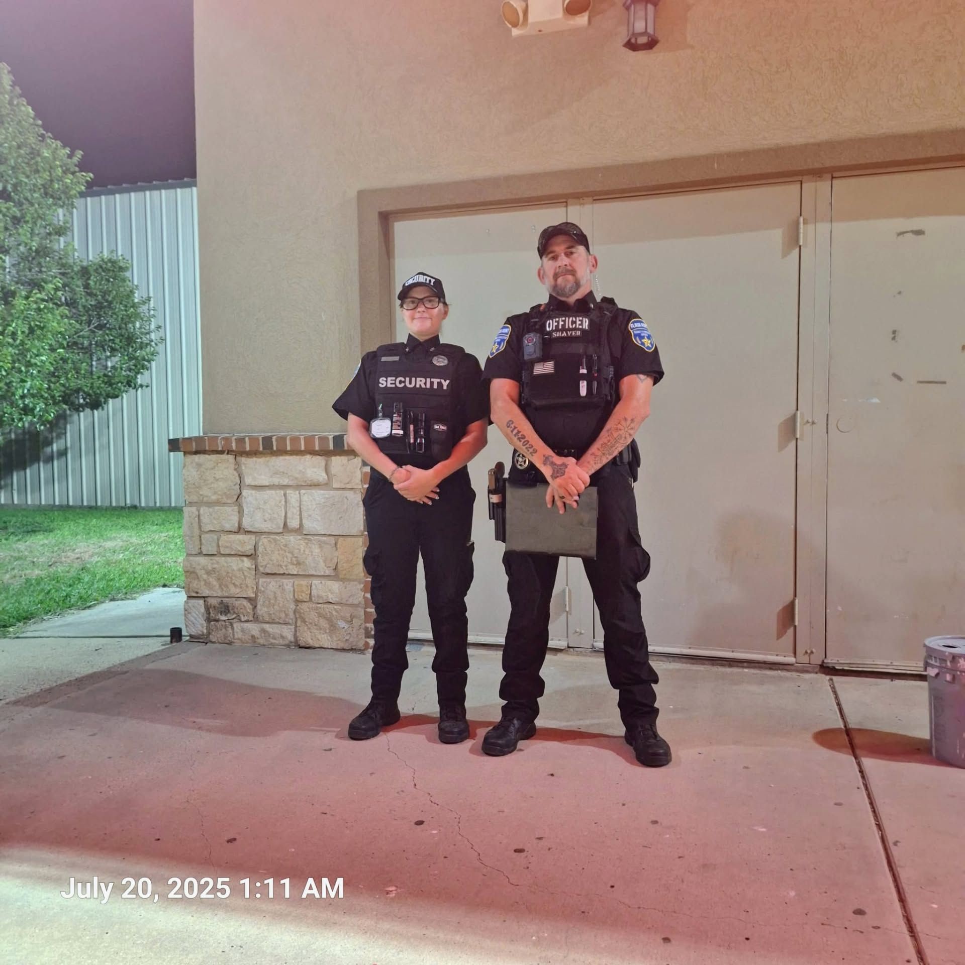 Two security officers in black uniforms stand outside a building at night.