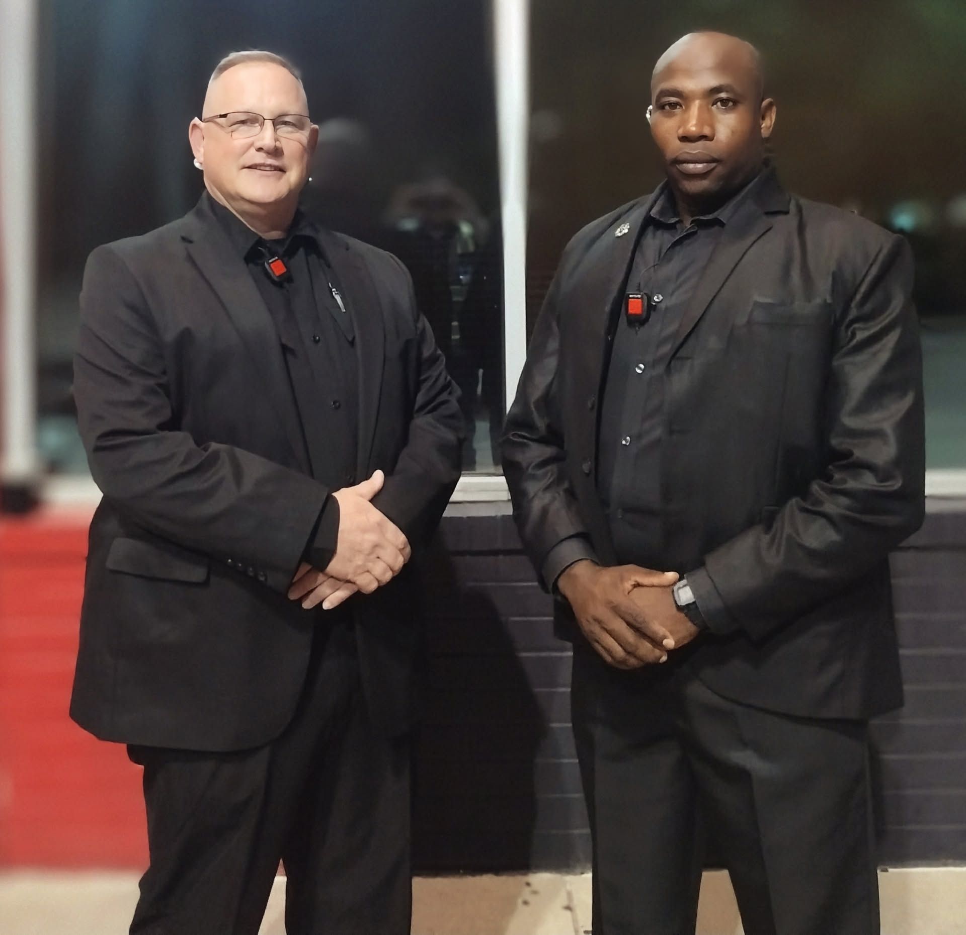 Two men in black suits standing on a red carpet, posing with hands clasped.