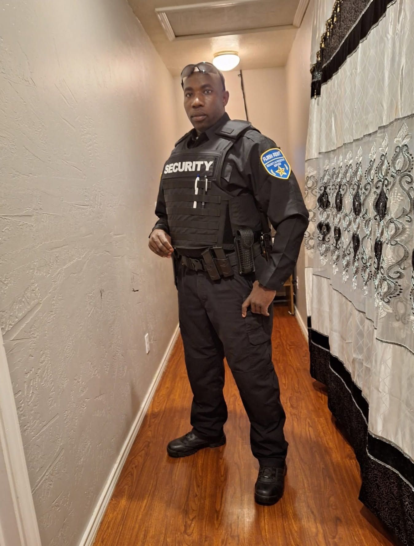 Security guard standing in a hallway, wearing a black uniform and vest.
