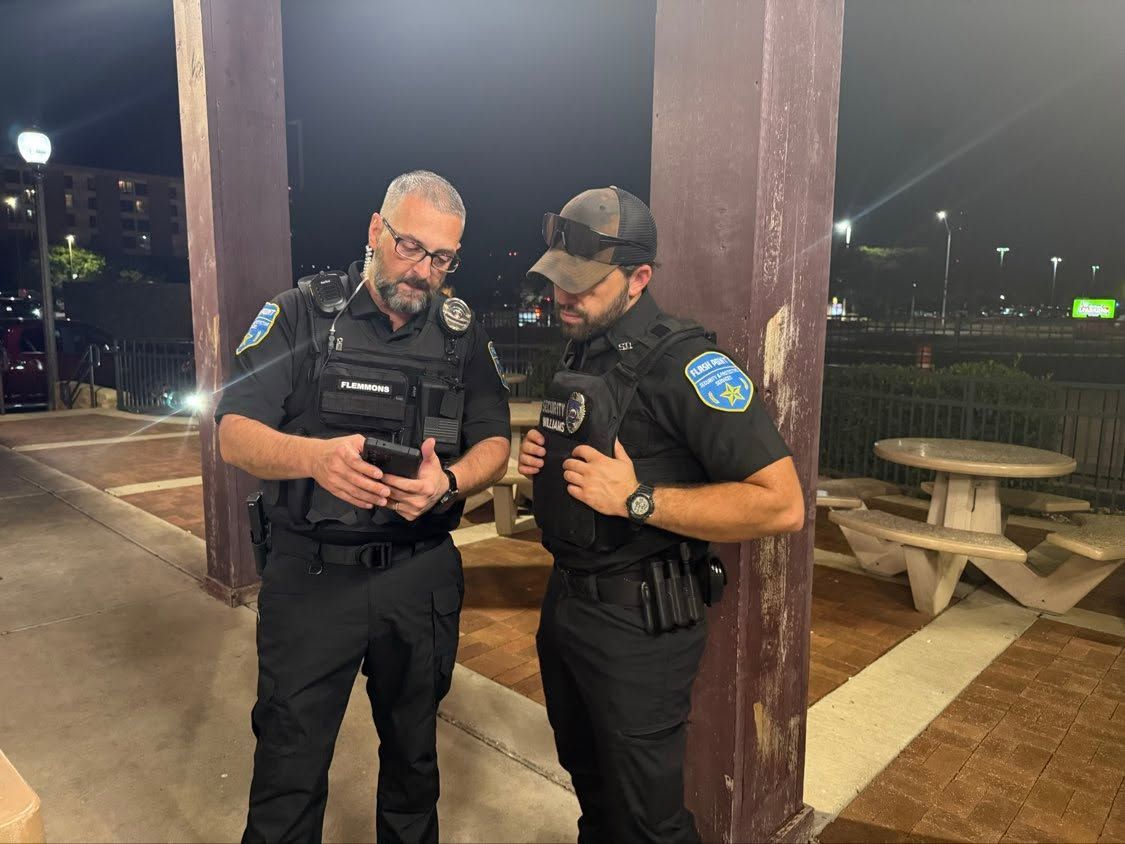 Two police officers in black uniforms stand under a lit pavilion, looking at a device together at night.