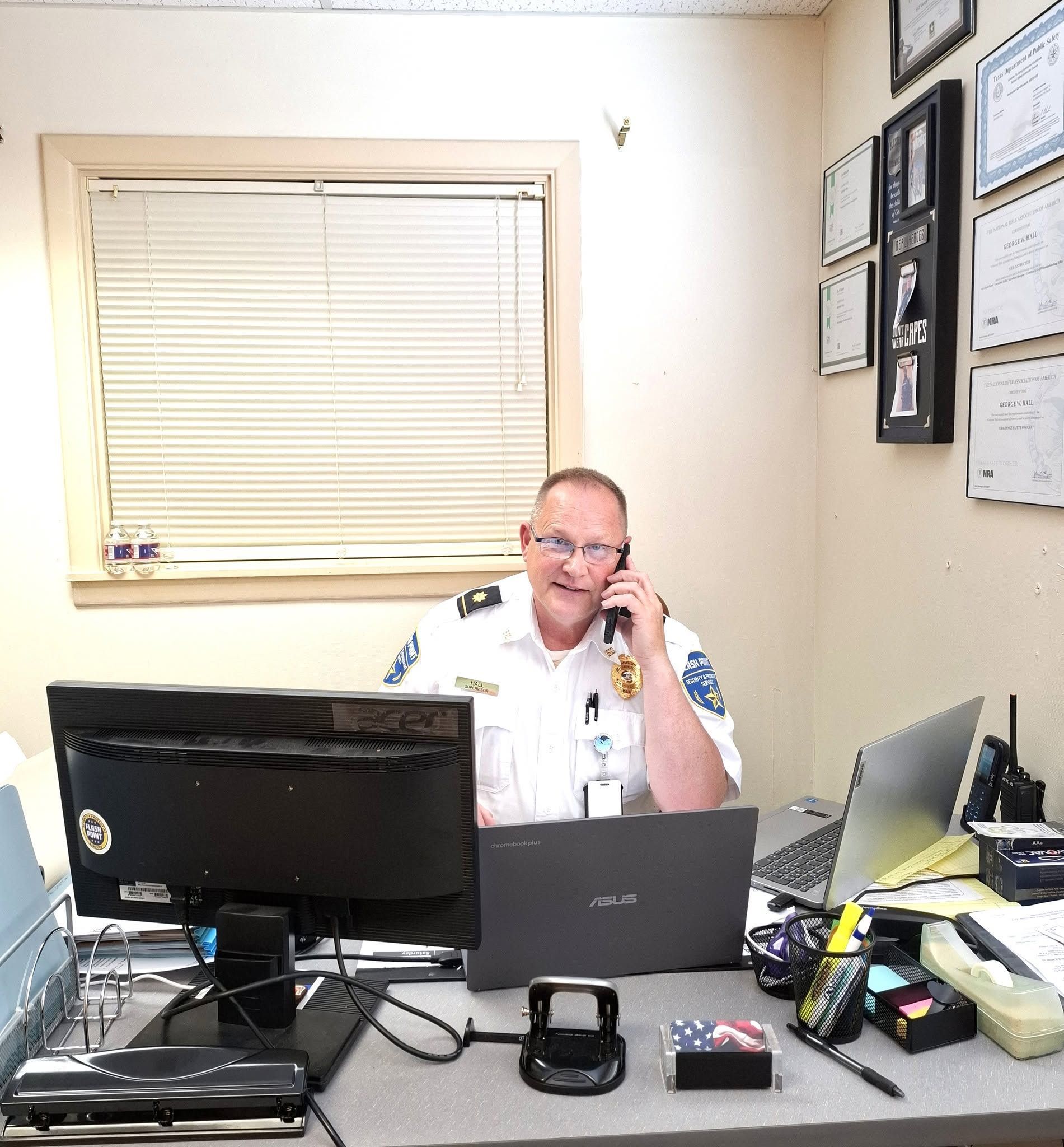 Man seated at office desk on phone, with computer monitors, papers, and framed documents on the wall.