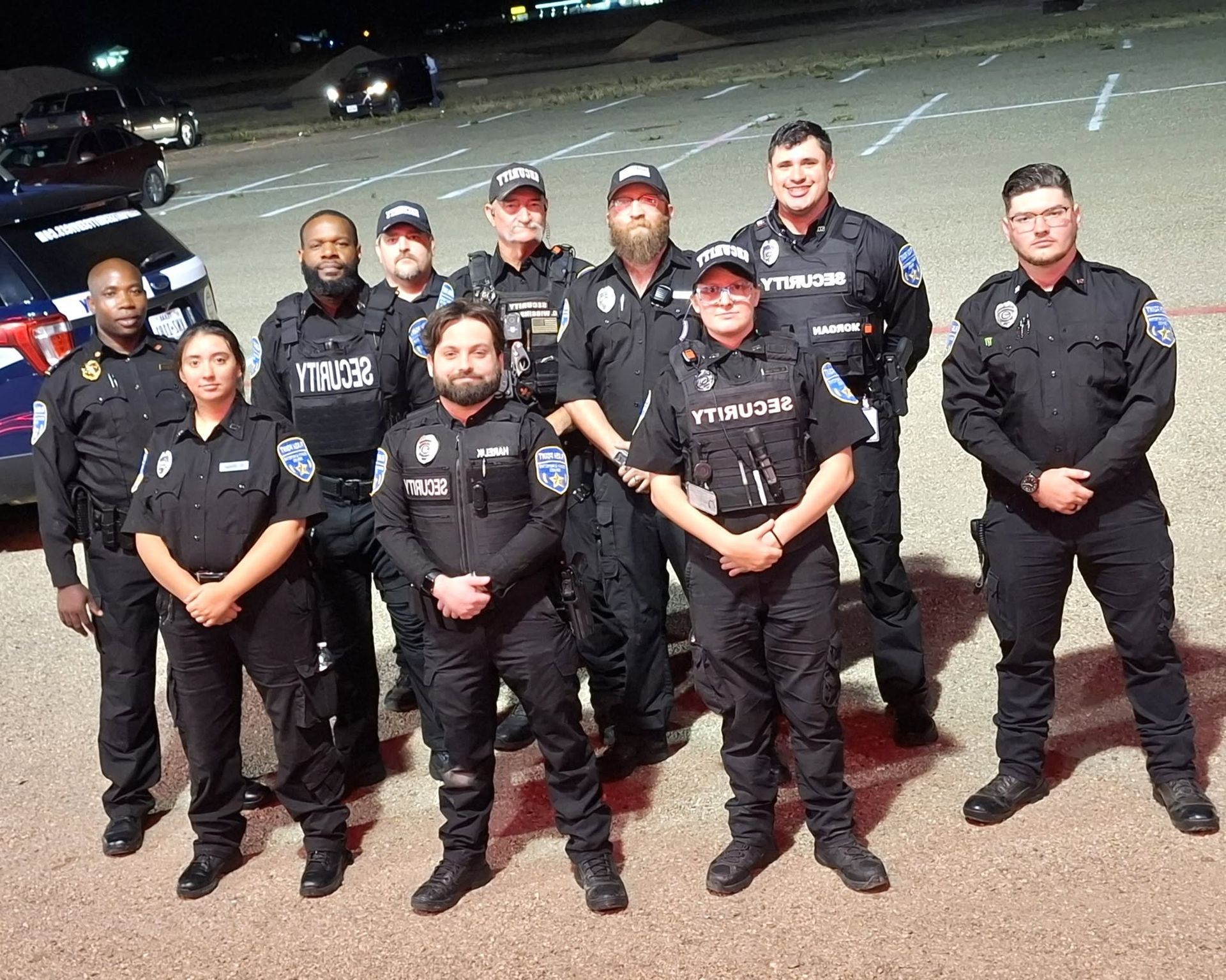 Police officers standing in formation outdoors at night, wearing black uniforms, with cars behind them.