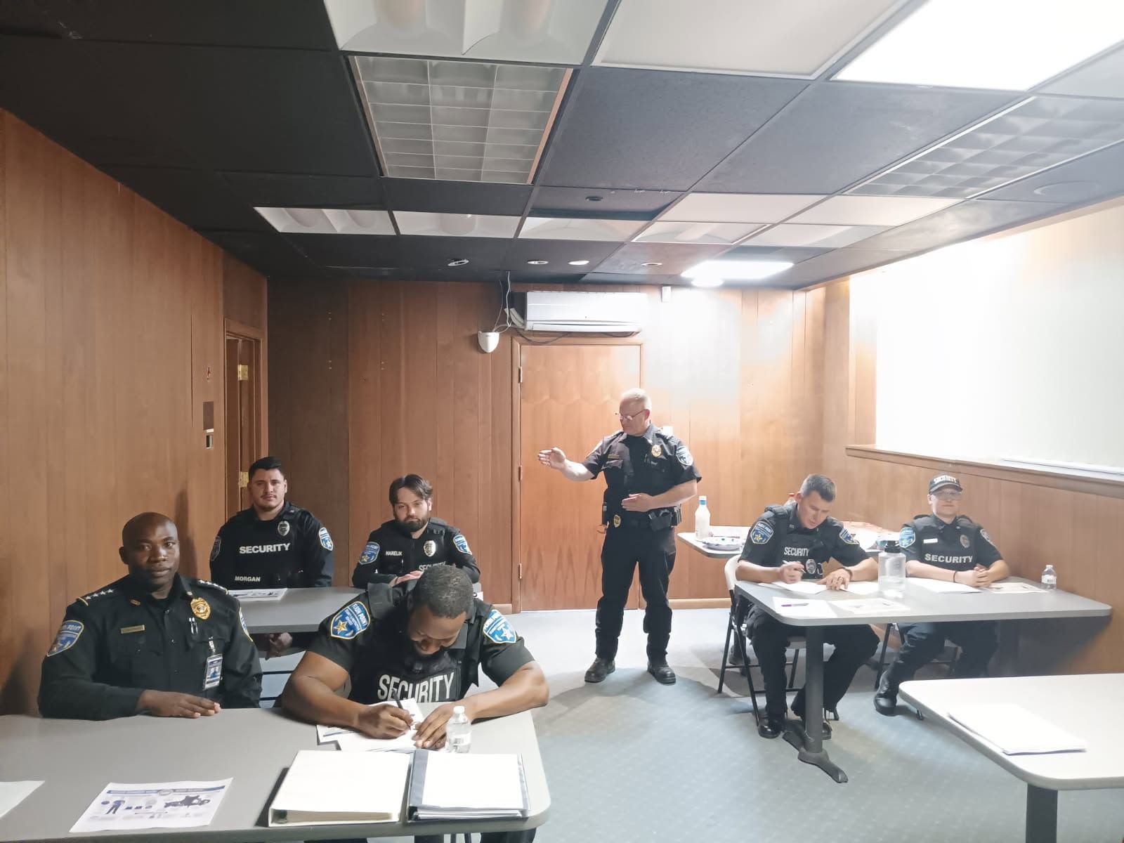 Police officers seated in a wood-paneled room, one standing and gesturing near a table.