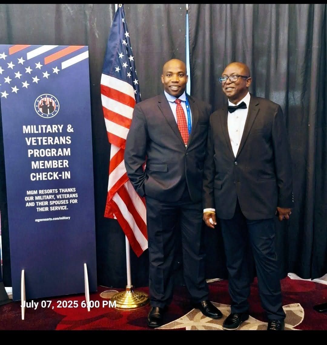 Two men in tuxedos standing beside a Military & Veterans Program check-in sign and U.S. flag.