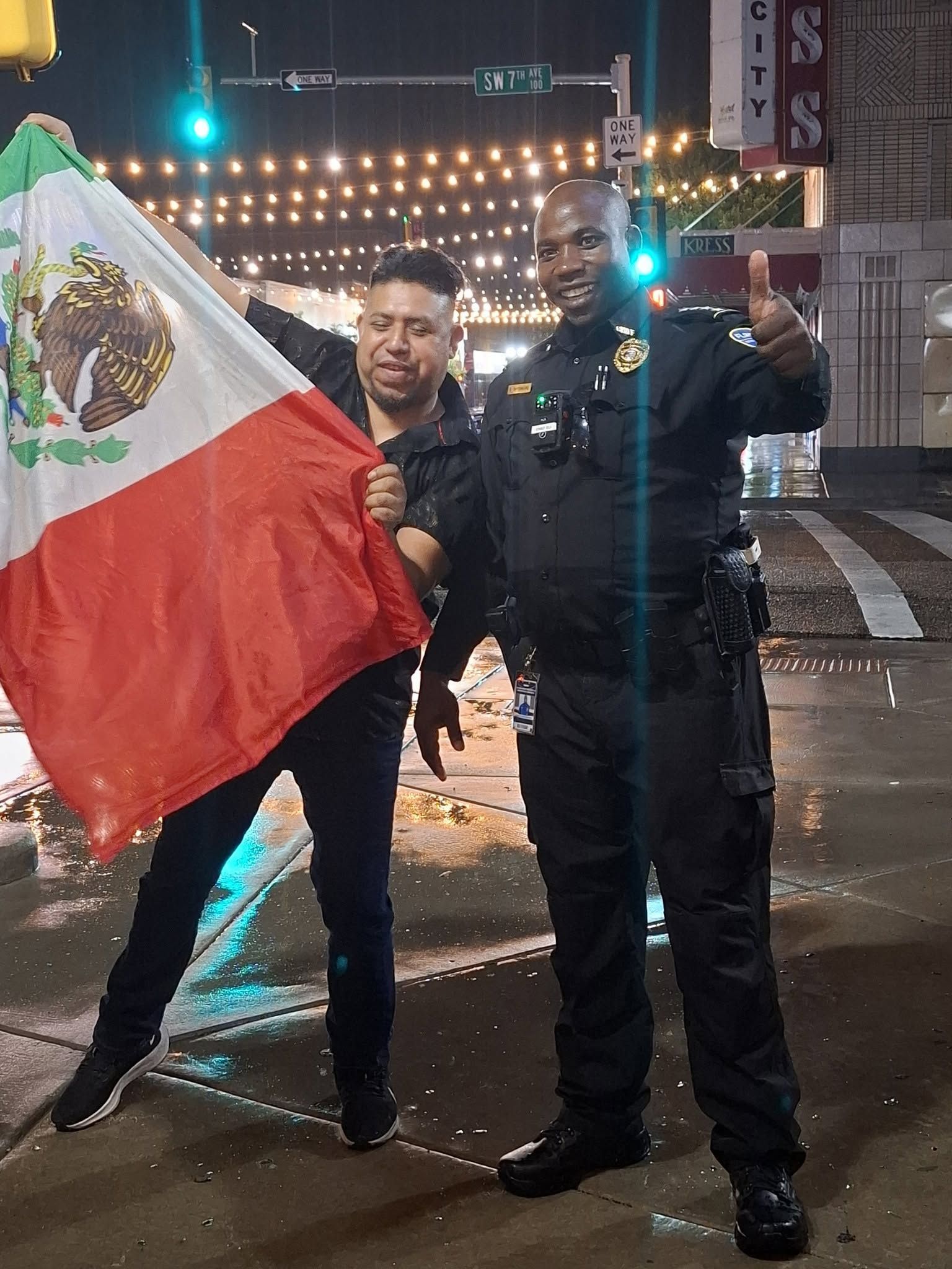 Two men posing on a wet city street at night, one holding a Mexican flag.
