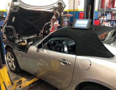Silver convertible car with hood open on a lift in a garage. A diagnostic tool sits on the folded convertible top.