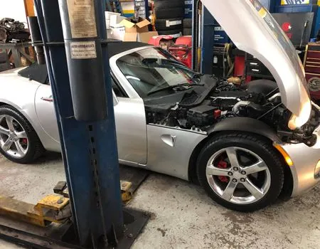 Silver sports car with hood open in a repair shop, showing engine bay and lifted on a car lift.