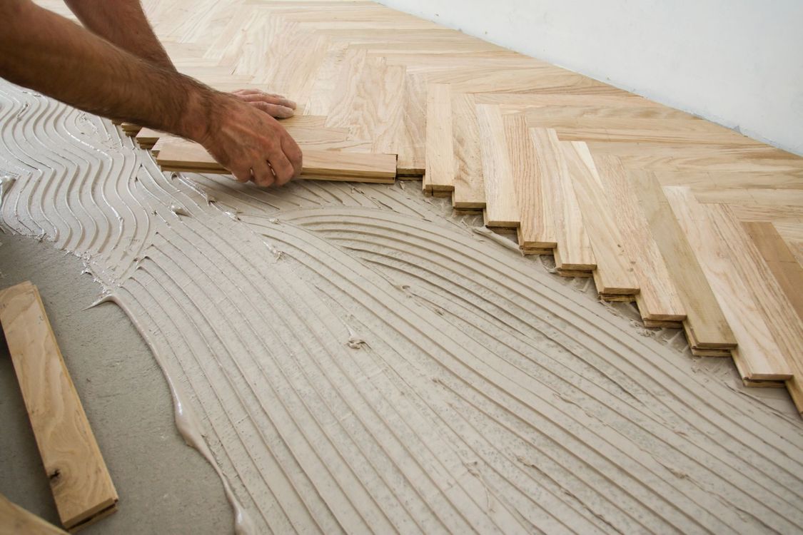 Mani che posano assi di legno chiaro per pavimenti, disposte a spina di pesce, su una base adesiva stesa con la cazzuola.