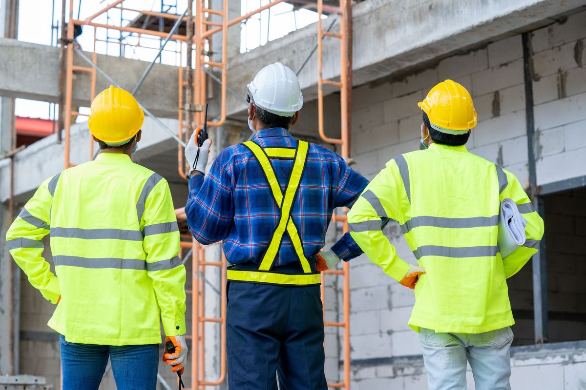 Tre operai edili, con indosso giubbotti ad alta visibilità e caschi protettivi, si trovano in un cantiere e guardano verso l'alto, verso le impalcature.