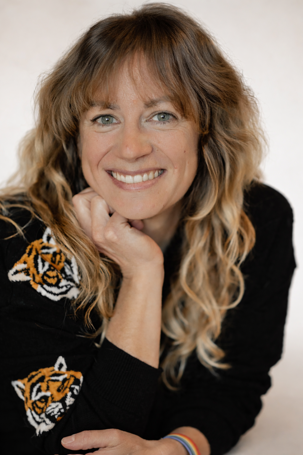 Smiling woman with long wavy hair resting her chin on her hand, wearing a black sweater with tiger designs.
