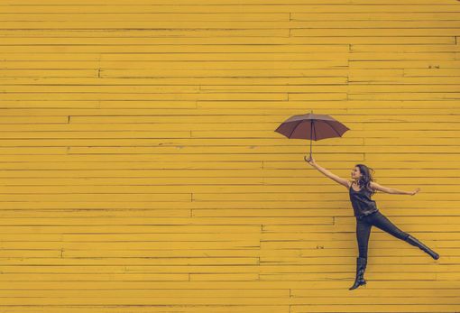 Person holding an umbrella, jumping beside a bright yellow wooden wall.