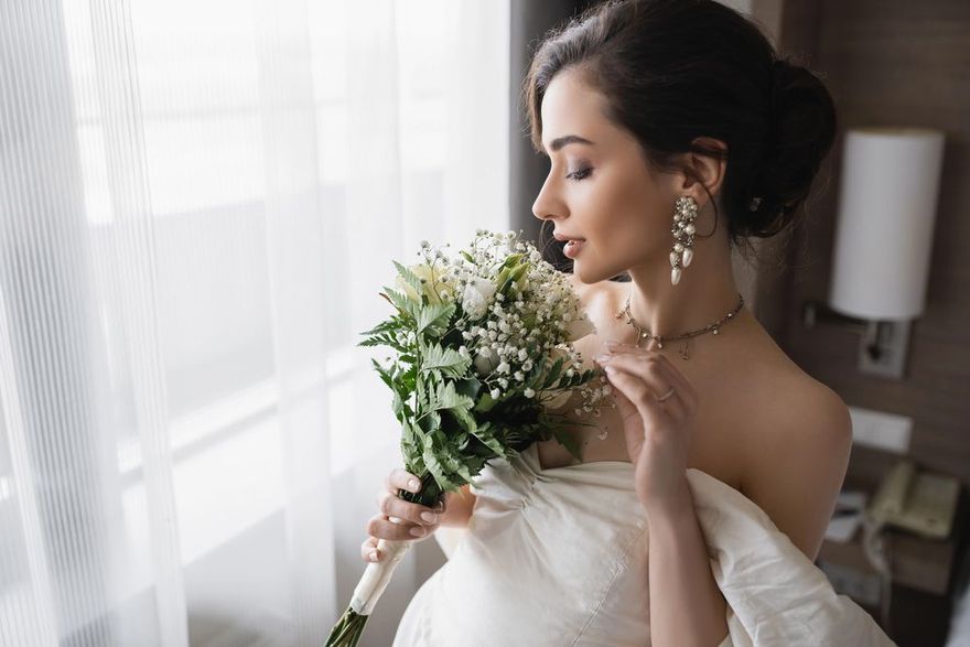 Una donna in vestaglia bianca tiene in mano un mazzo di fiori accanto a una finestra, in una camera da letto illuminata da una luce soffusa.