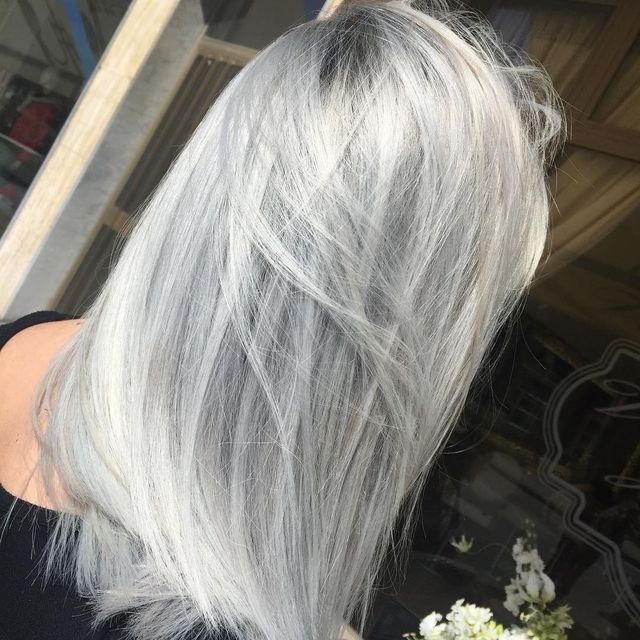 Vista laterale di una persona con lunghi capelli bianco-argento, in piedi all'aperto vicino a una finestra.