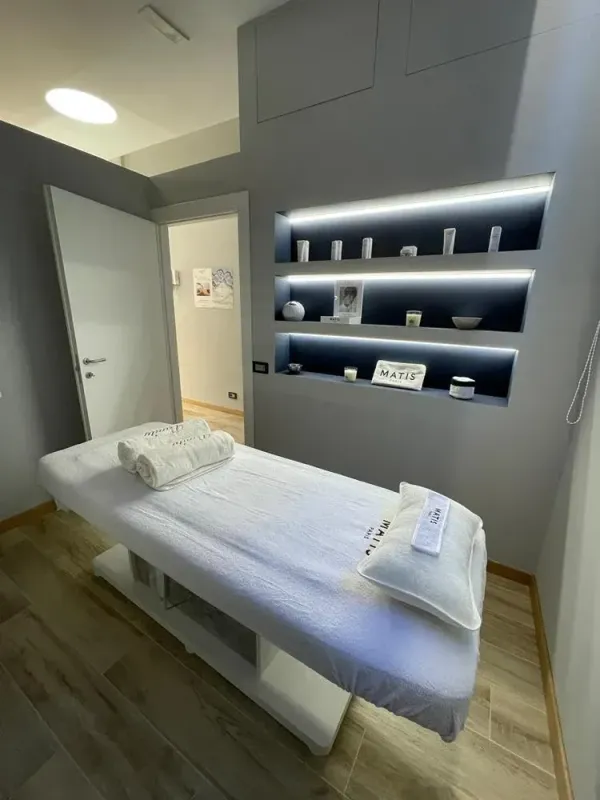 Moderna sala per trattamenti spa con lettino da massaggio, asciugamani piegati e mensole a parete illuminate con prodotti