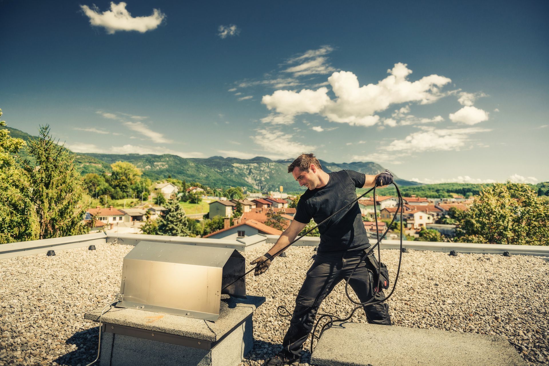 A worker in a black uniform, performing chimney sweep services, cleaning a chimney on top of a roof.