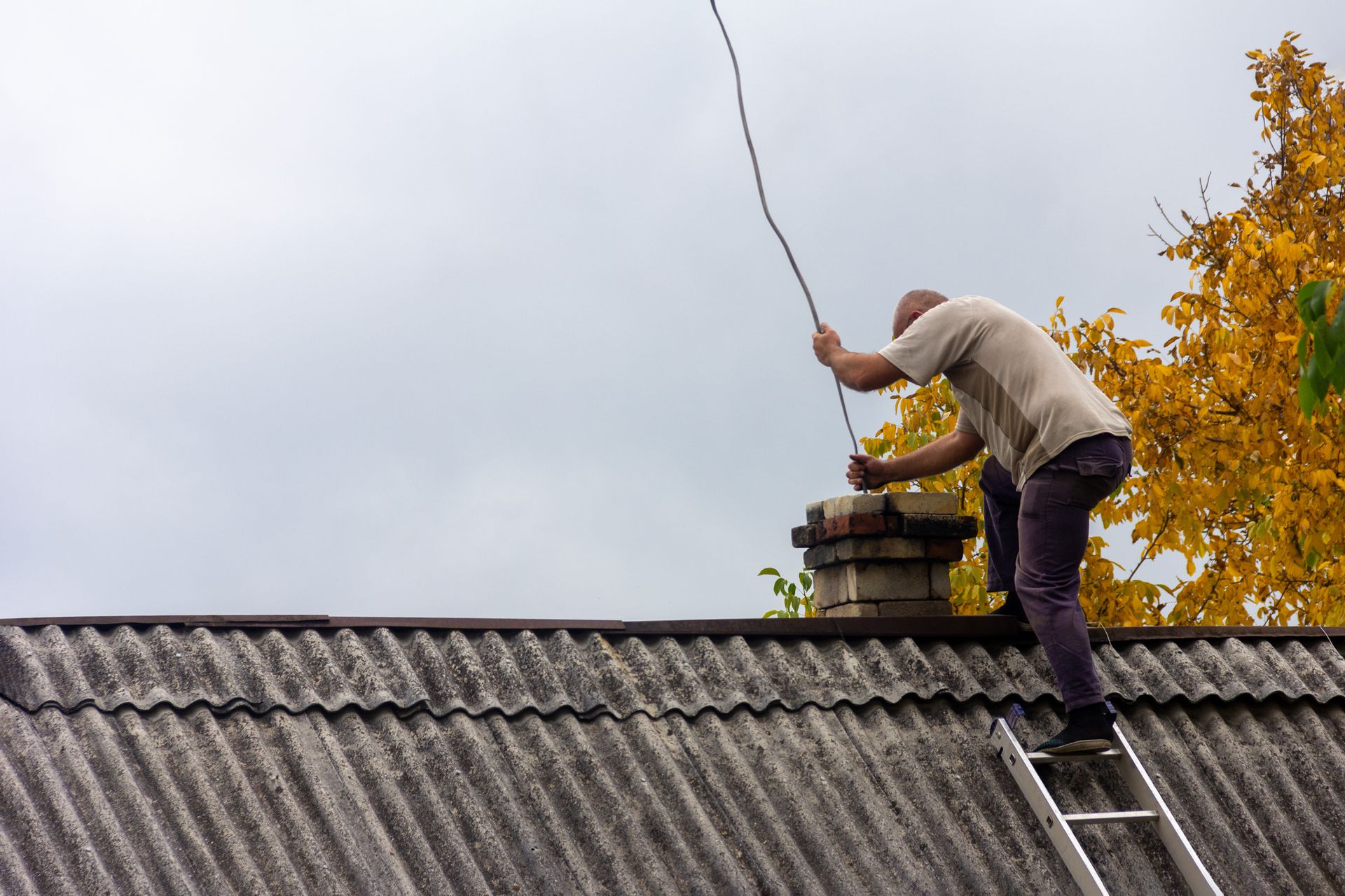 Man on ladder near chimney, overcast sky. Reaching for a wire on roof.
