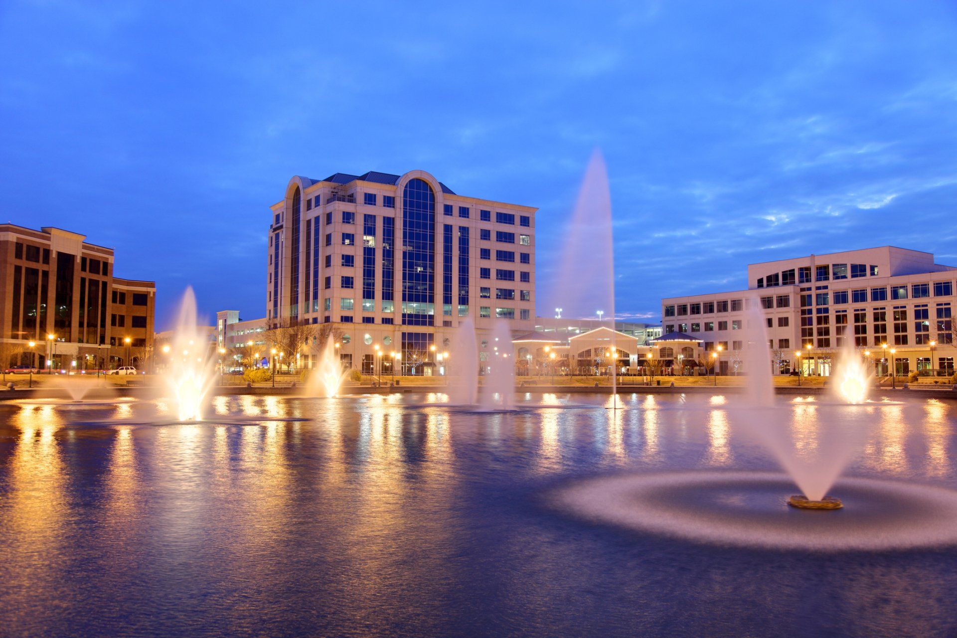 Fountains spray water in a lake in front of illuminated buildings under a twilight sky.