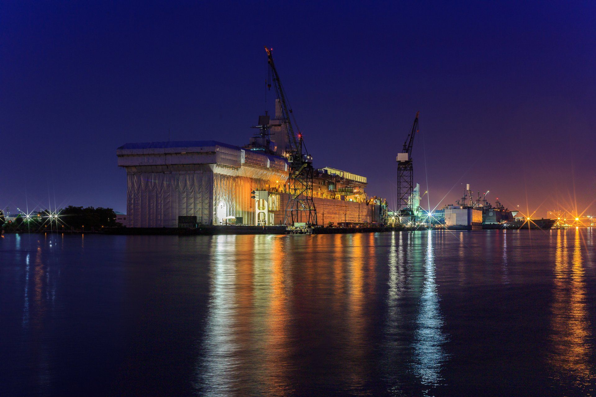 Night view of a lit shipyard with large cranes and a docked vessel, reflected in dark water against a deep blue sky.