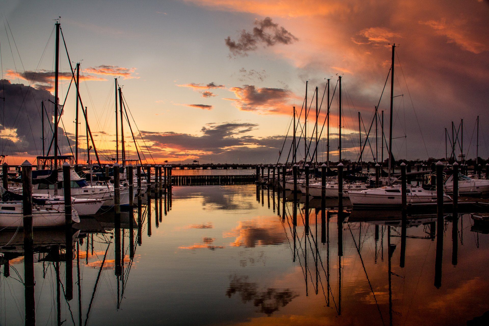 Sailboats docked at a marina reflect in the still water during a vibrant sunset, with orange and pink hues in the sky.