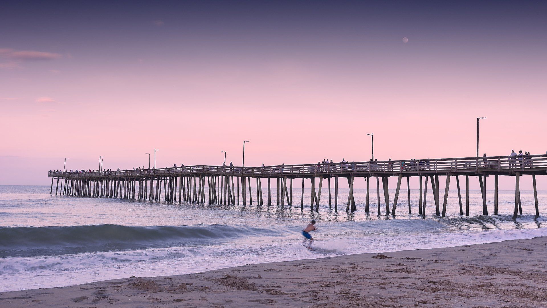 A wooden pier extends over ocean waves at dusk. A person runs in the shallow water, bathed in pink and purple light.