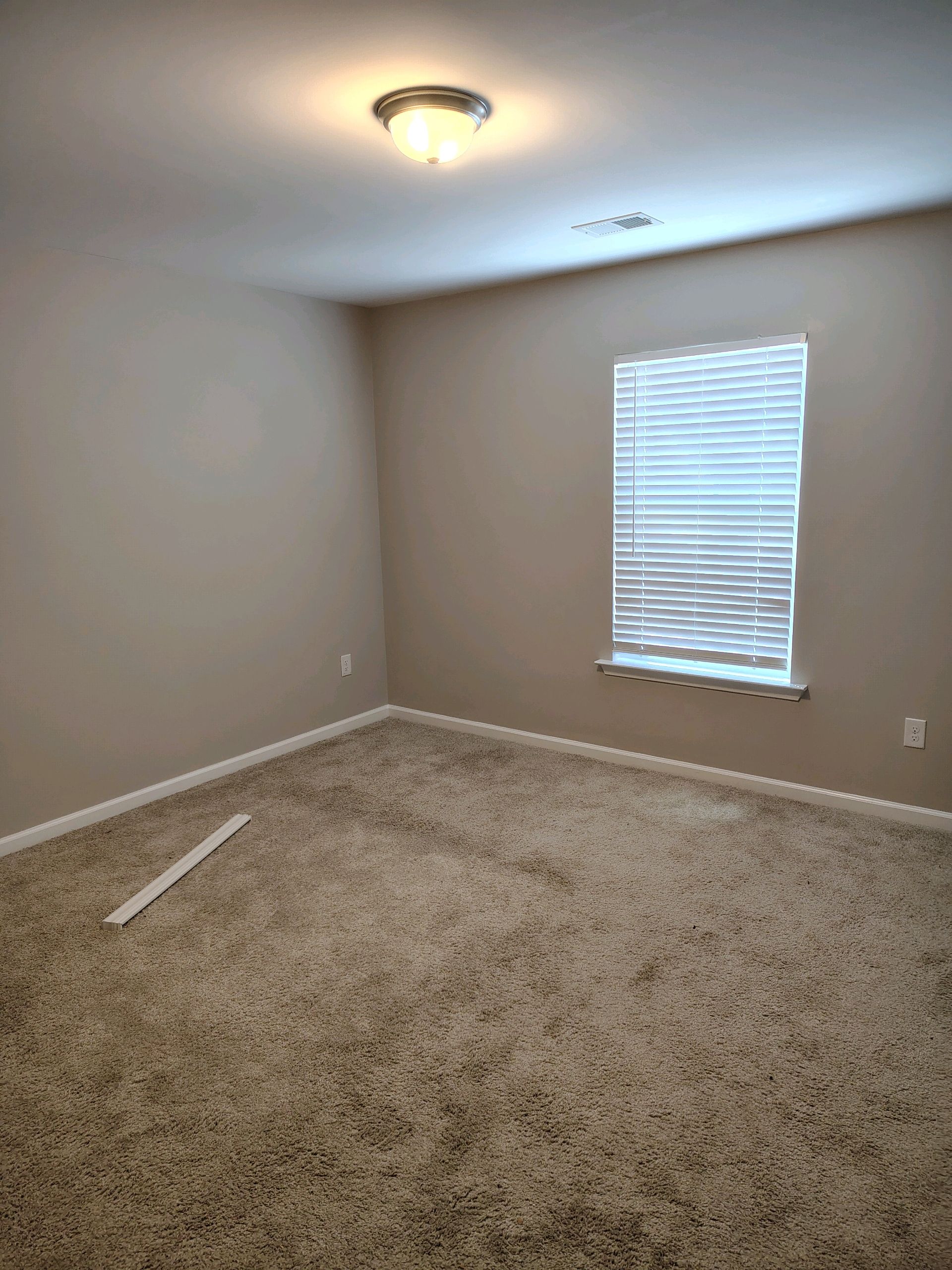 Empty beige bedroom with carpet, window blinds, and a ceiling light.