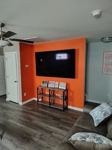 Living room with wall-mounted TV on orange accent wall, ceiling fan, and sectional sofa.