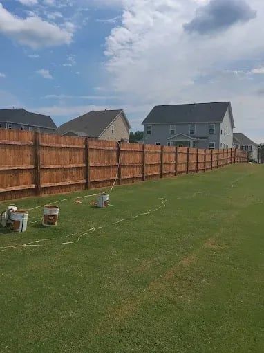 Long backyard with green lawn, wooden fence, and houses under a cloudy sky