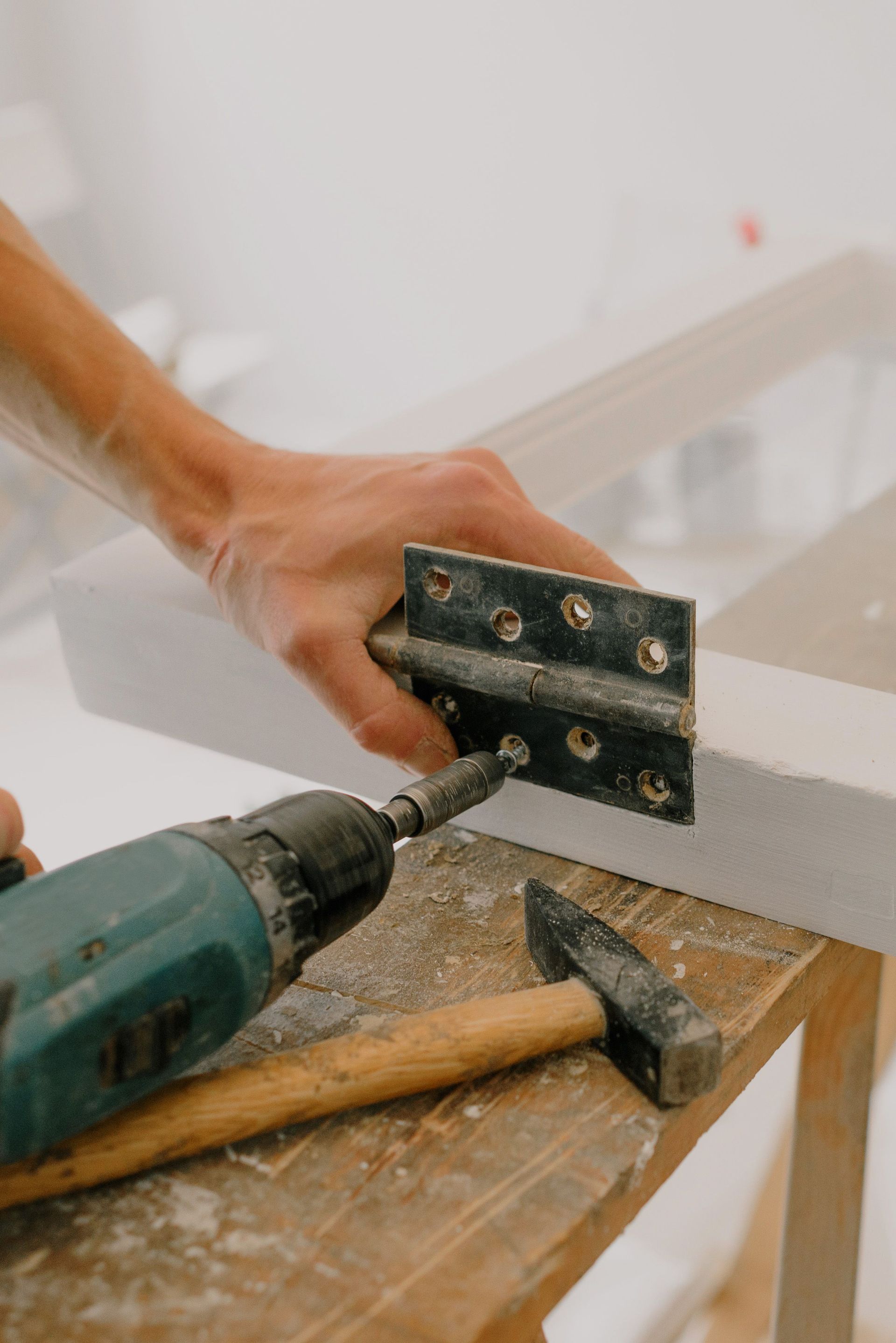 Hand holding a drill near a metal hinge, with a hammer on a workbench