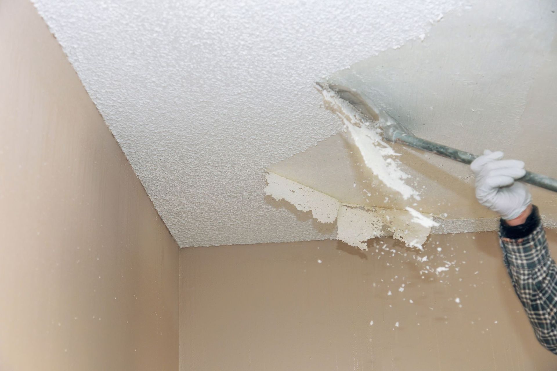 Person scraping damaged ceiling drywall with a tool, beige room with debris falling