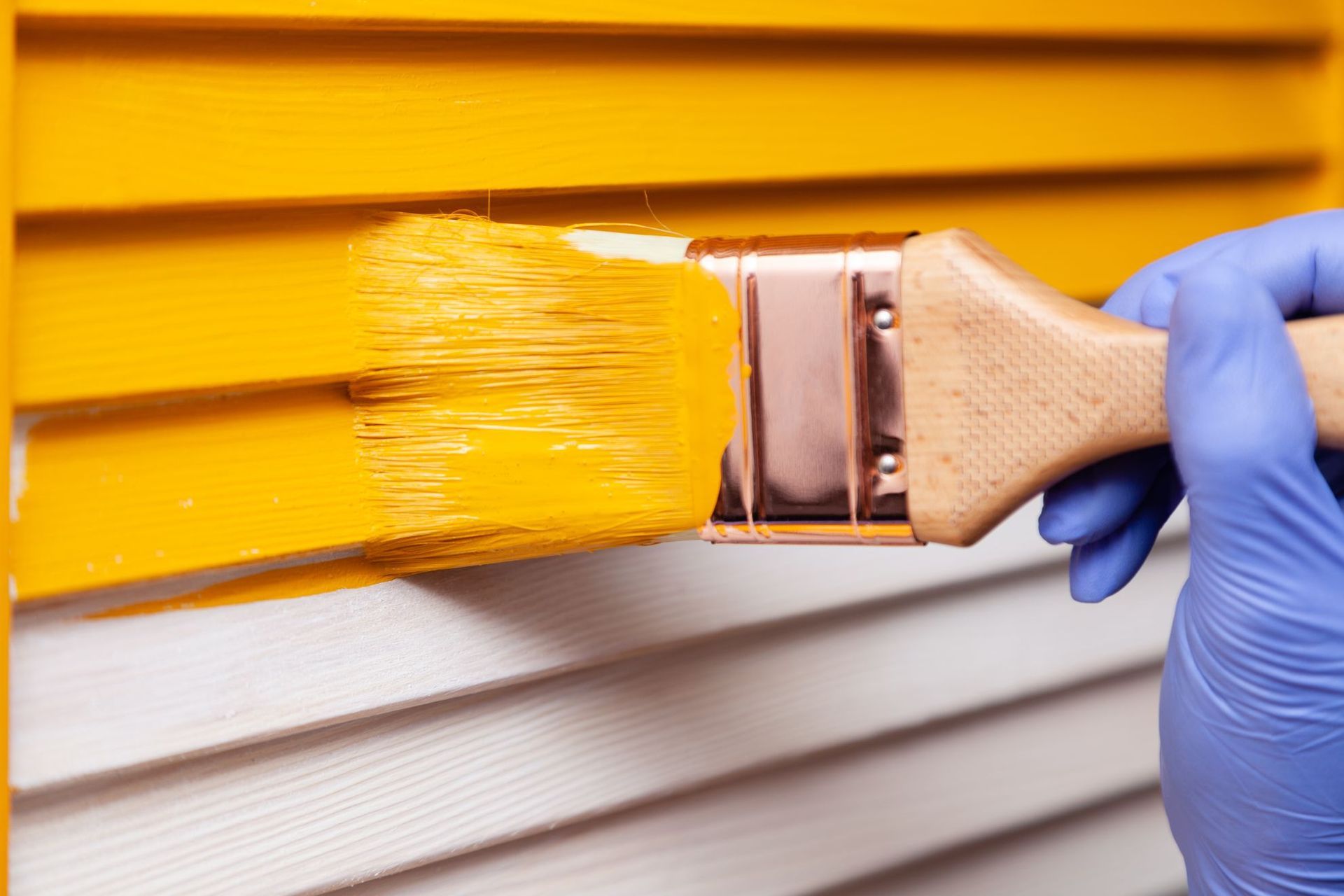 Gloved hand painting a white wall with bright yellow paint using a brush