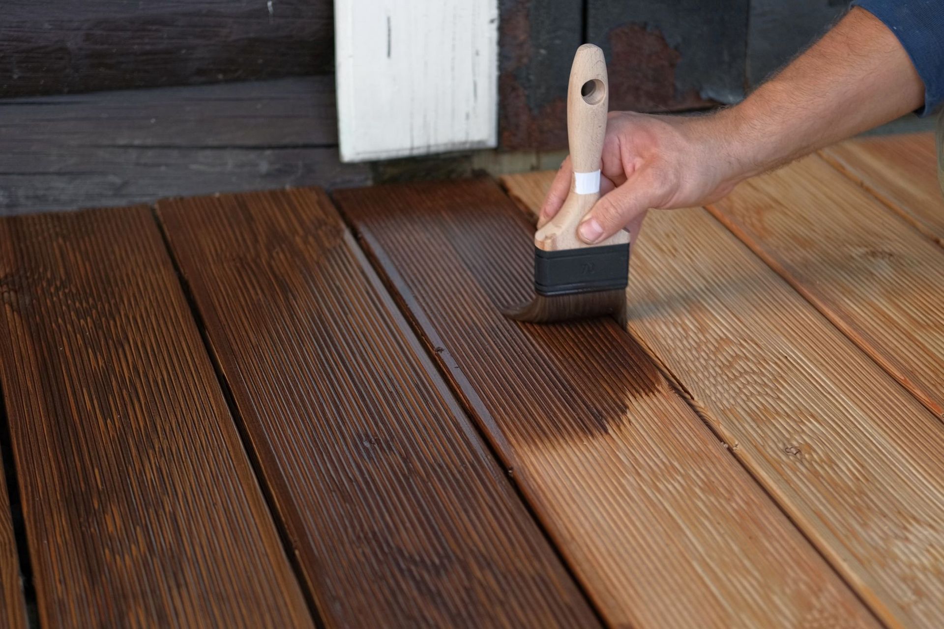 Hand brushing dark stain onto wooden deck boards beside a painted white surface