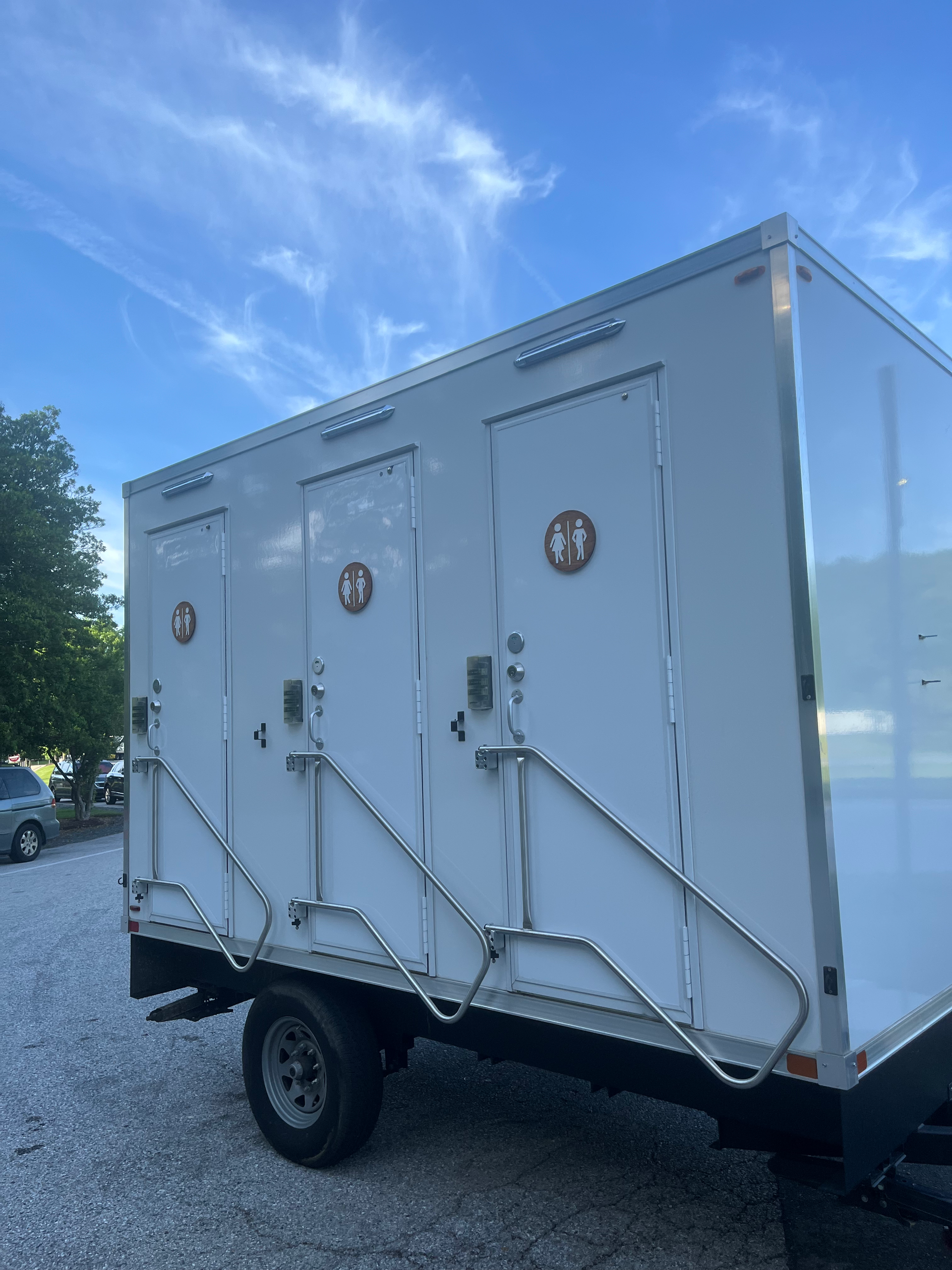 A white trailer with three toilets on it is parked in a gravel lot.