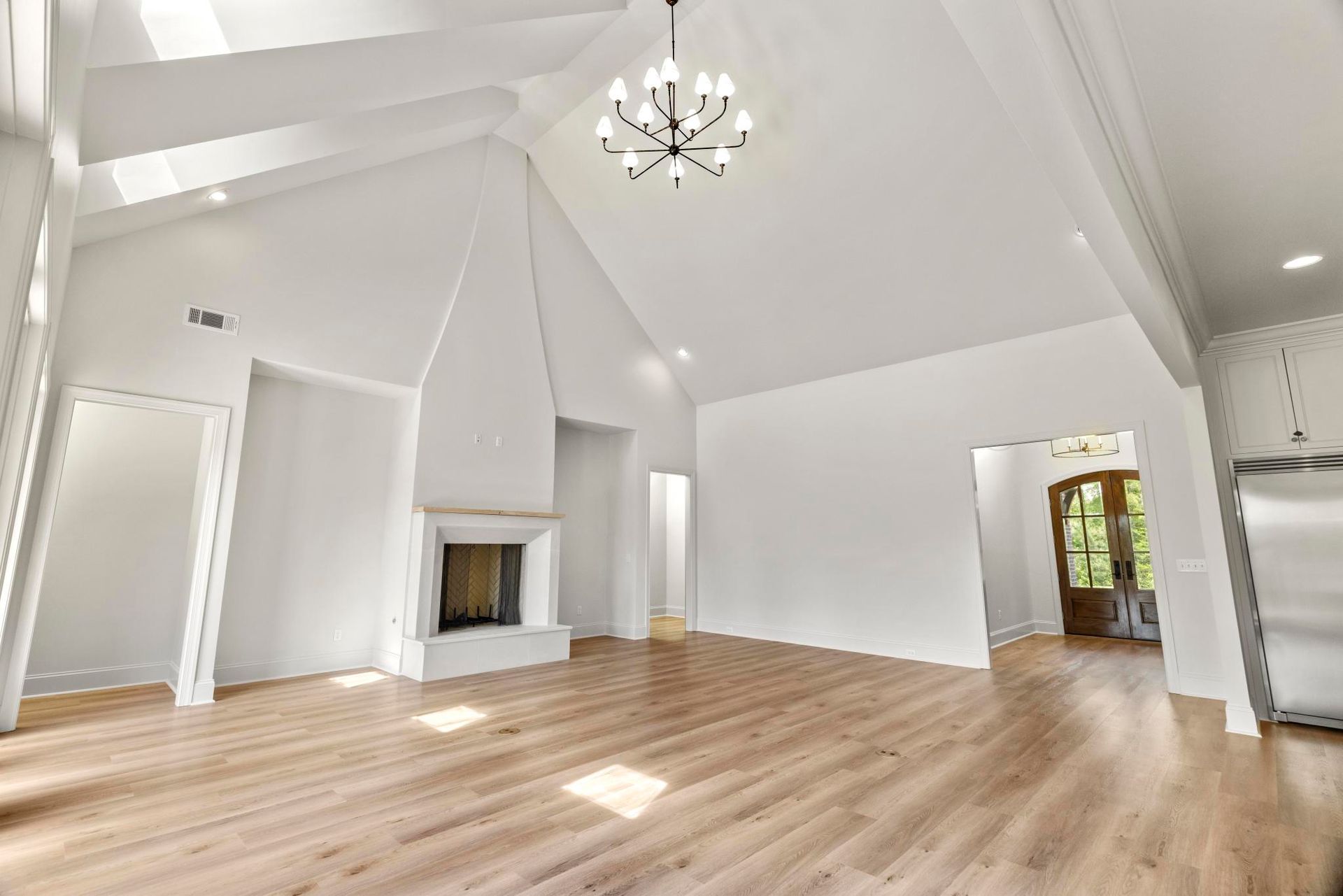 Spacious empty living room with white vaulted ceilings, fireplace, and light wood floors