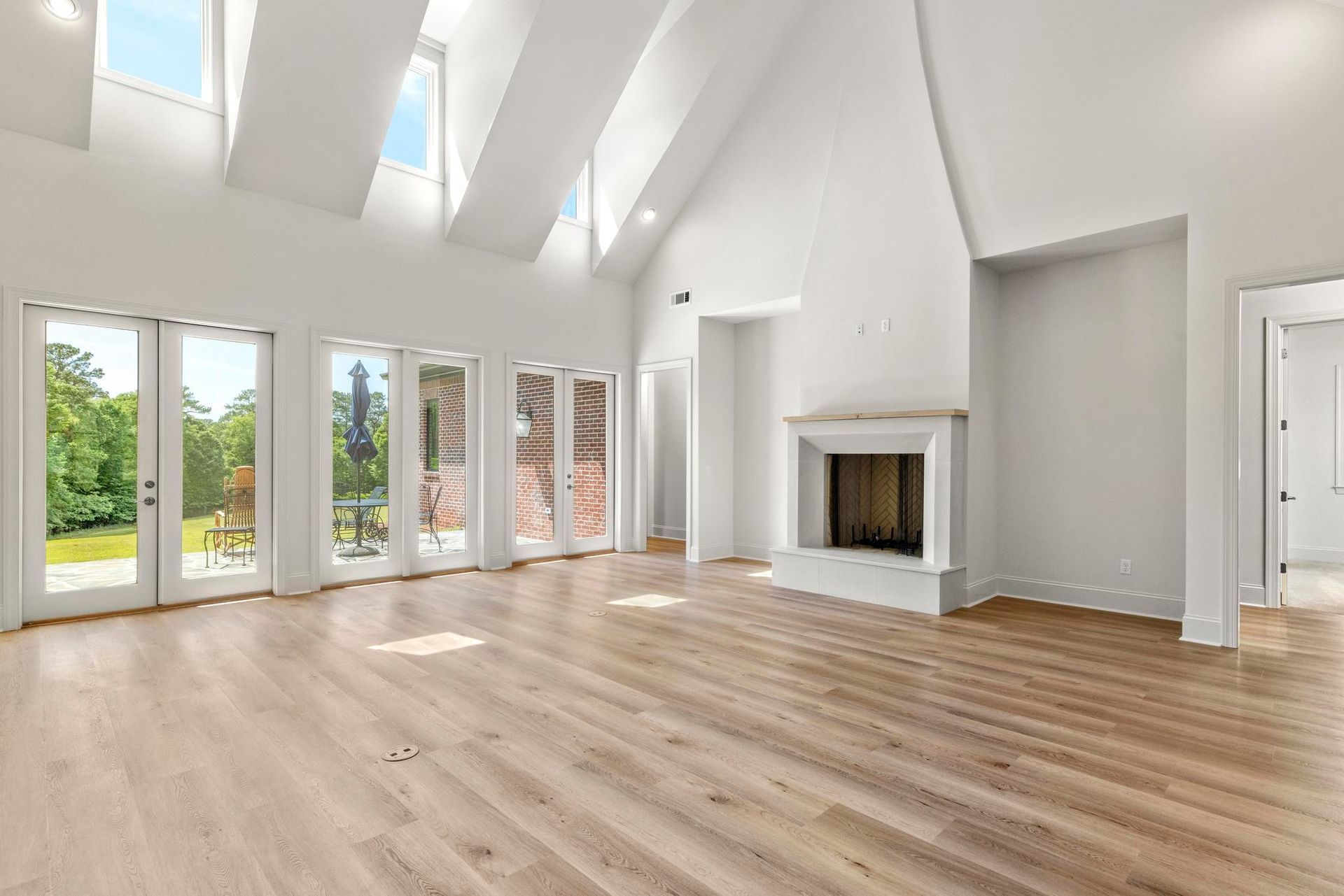 Bright empty living room with vaulted white ceiling, large windows, wood floors, and a fireplace