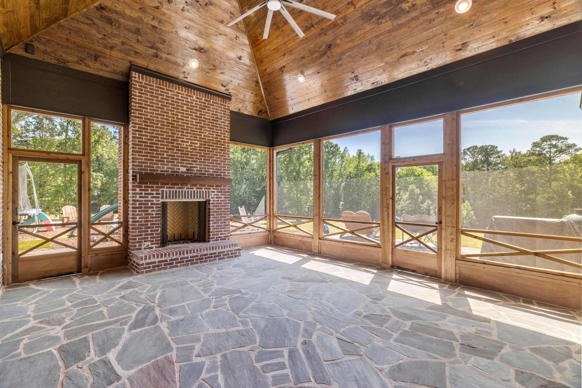 Spacious sunroom with stone floor, brick fireplace, wood ceiling, and large windows overlooking the outdoors