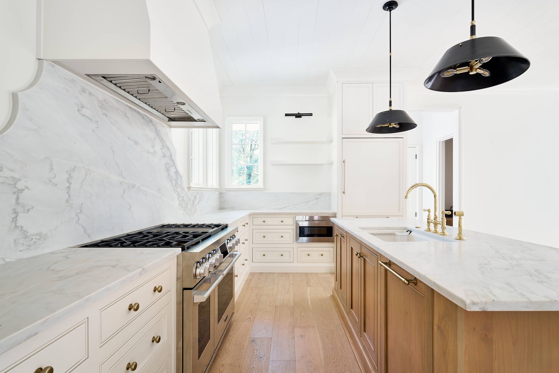 Bright modern white kitchen with marble counters, wood floors, black pendant lights, and gold faucet