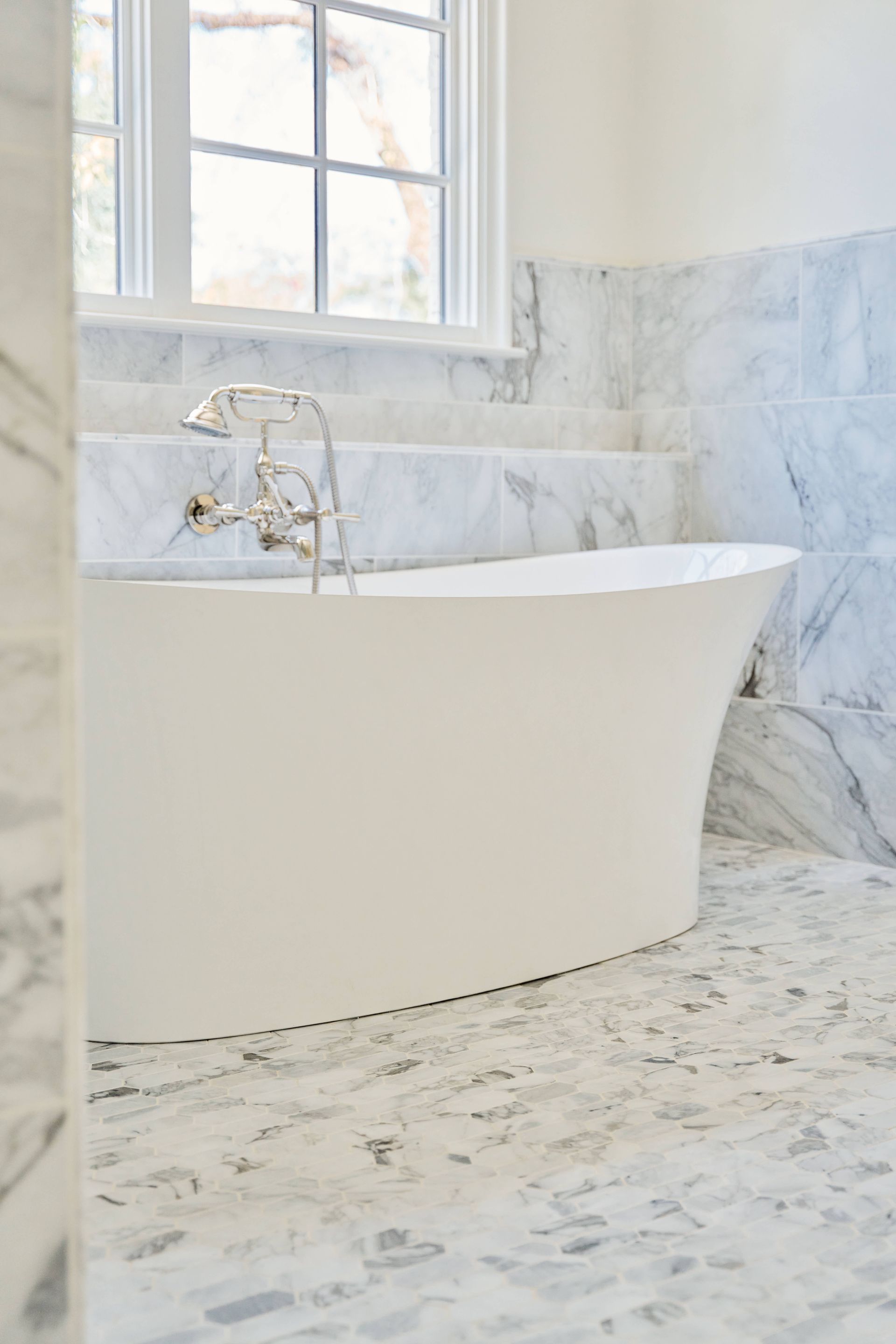 Bright white freestanding bathtub in a marble-tiled bathroom by a window