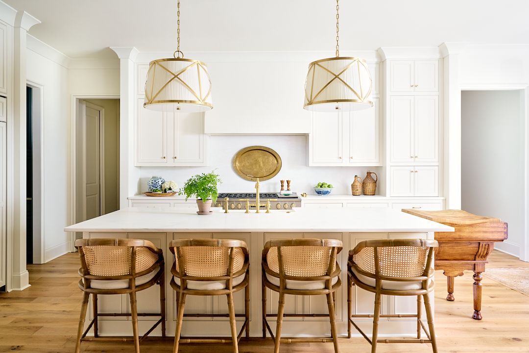 Bright white kitchen with island, woven bar stools, brass pendant lights, and a wooden table by the side.
