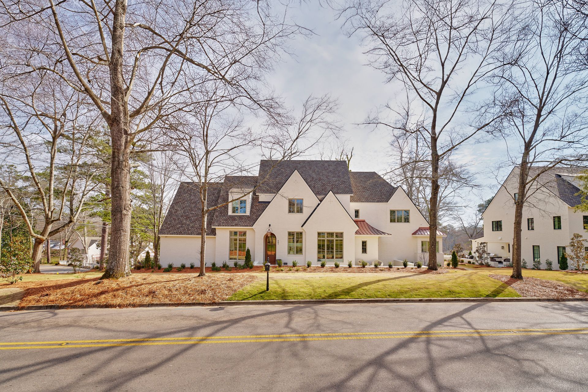 Large white house with dark roof behind bare trees, viewed from a quiet street on a sunny day
